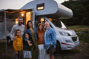 Um retrato de uma família de várias gerações olhando para a câmera e sorrindoao ar livre ao anoitecer, caravana viagem de férias.