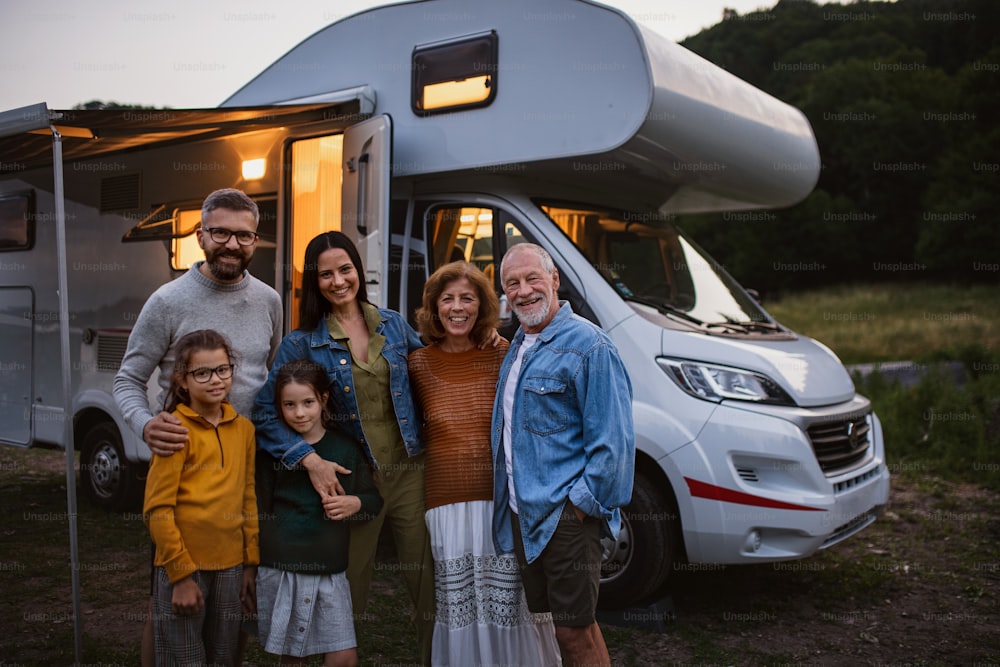 A portrait multi-generation family looking at camera and smilingoutdoors at dusk, caravan holiday trip.