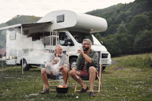 年配の父親を持つ成熟した男性が、屋外のキャンプ場で話し、キャラバン休暇旅行でバーベキューをする。