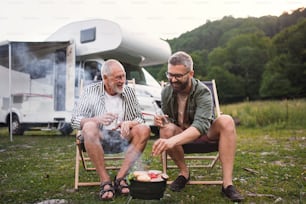 A mature man with senior father talking at campsite outdoors, barbecue on caravan holiday trip.