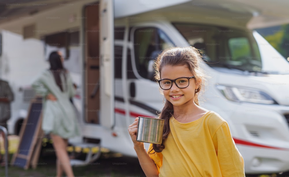 A happy small girl standing outdoors and looking at camera by caravan car, family holiday trip.