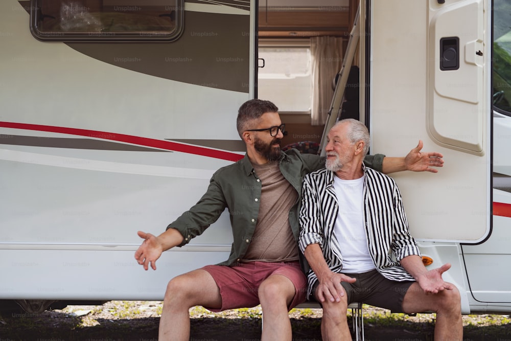 Cheerful mature man with senior father sitting by car outdoors, fun on caravan holiday trip.