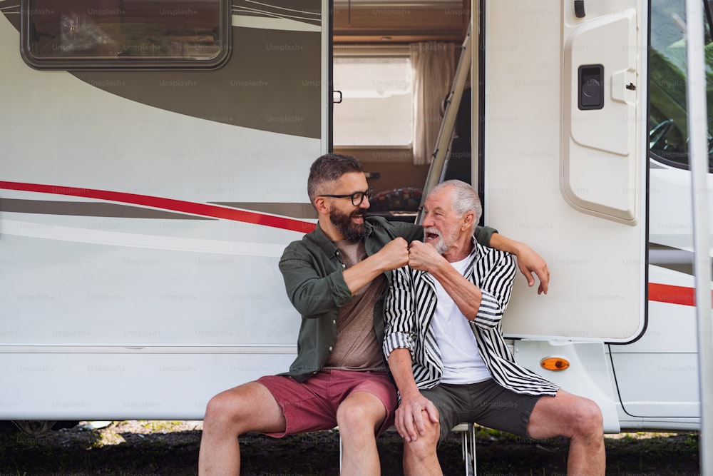 Cheerful mature man with senior father sitting by car outdoors, fun on caravan holiday trip.