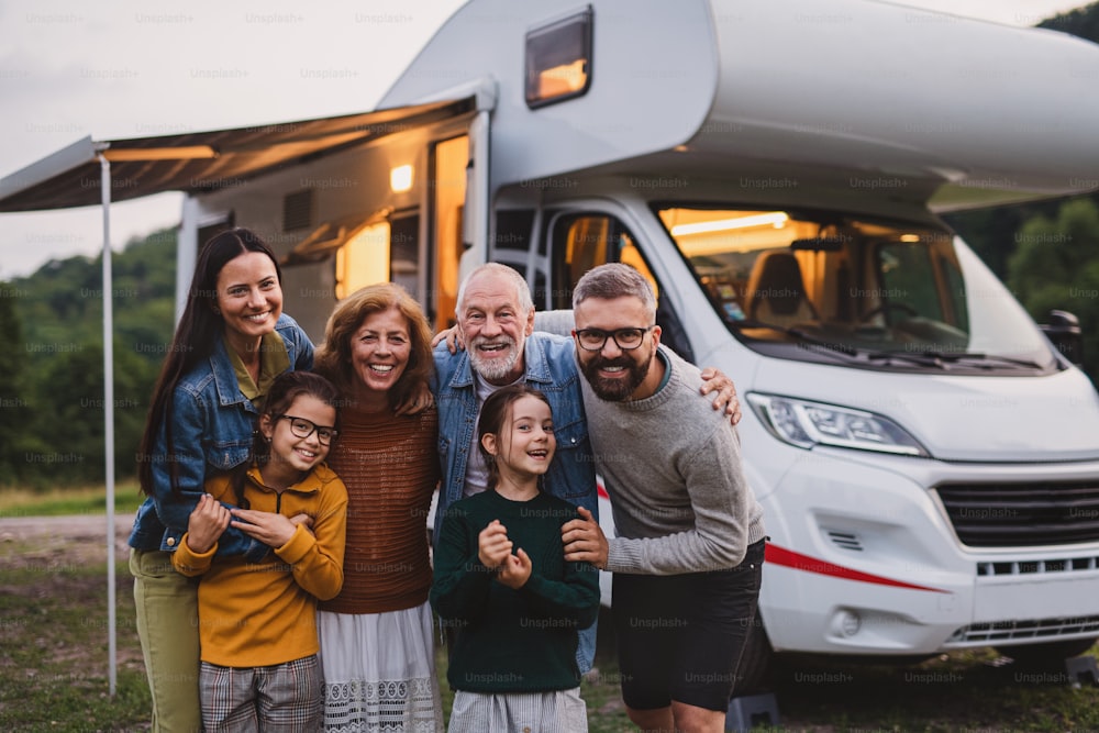 A multi-generation family looking at camera outdoors at dusk, caravan holiday trip.