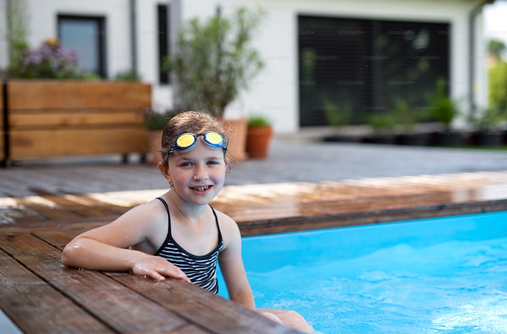 뒤뜰에서 고글을 쓴 작은 소녀가 수영장에 앉아 카메라를 보고 있다.