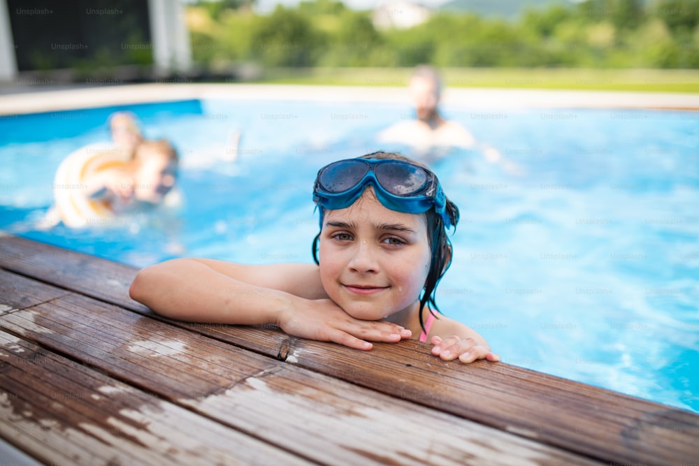 뒤뜰에서 고글을 쓴 작은 소녀가 수영장에 앉아 카메라를 보고 있다.