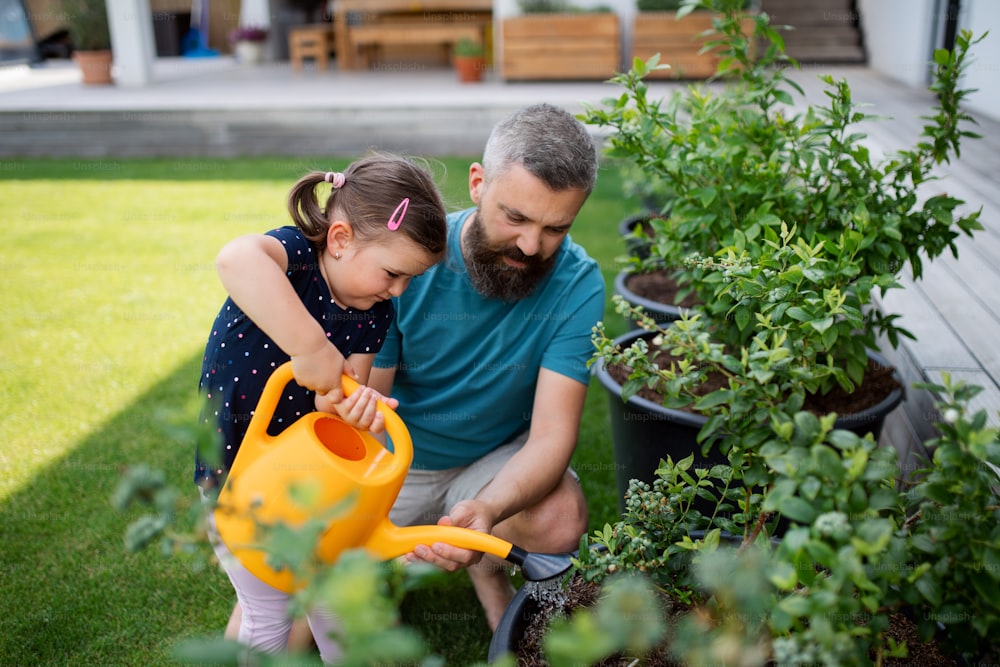 A father and small daughter with watering can outdoors in tha backyard, looking at camera.