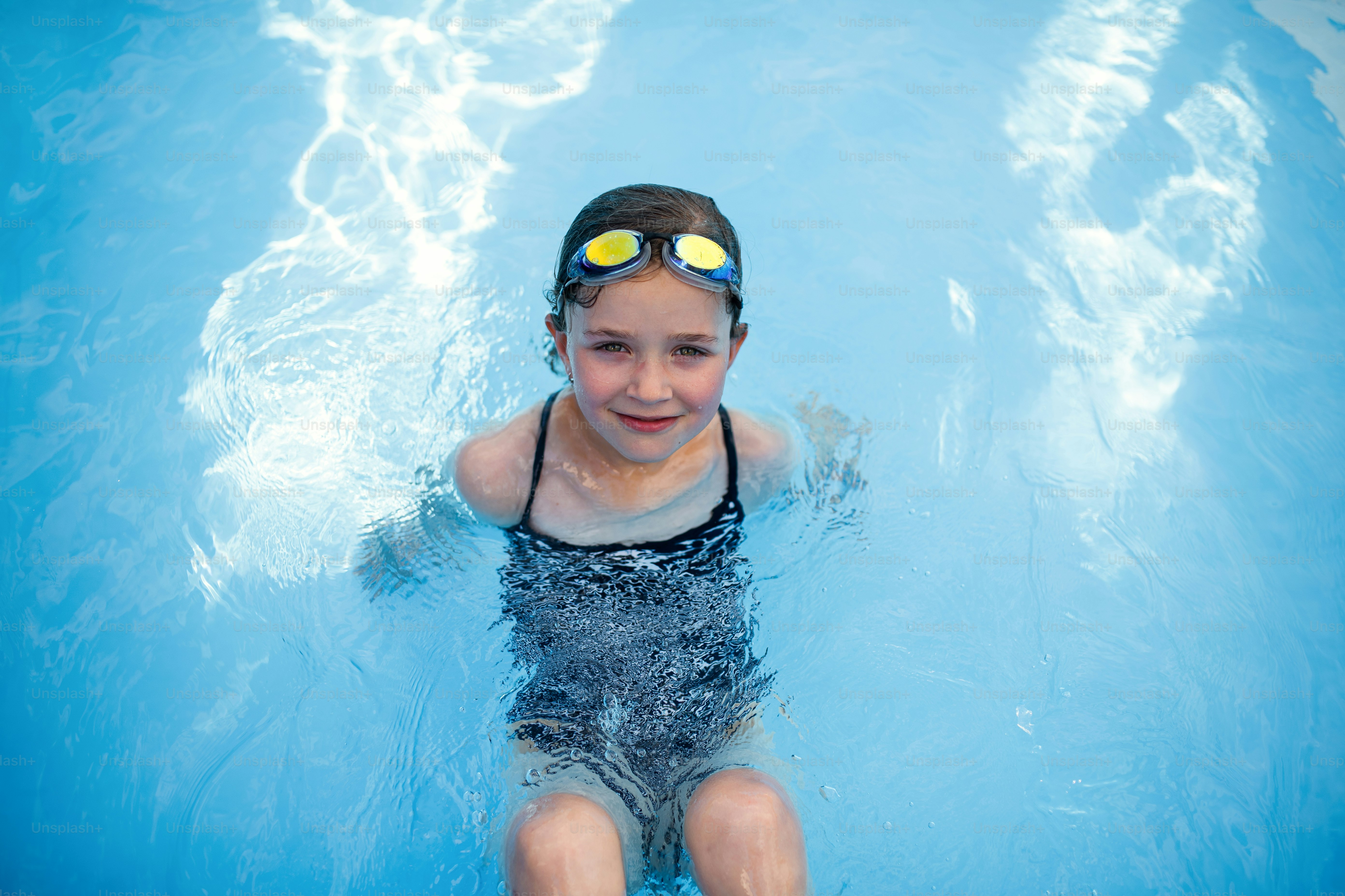 A top view of small girl outdoors in the backyard, sitting in swimming pool and looking at camera.