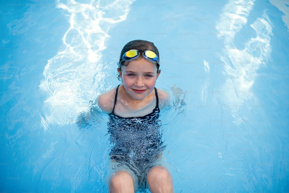 A top view of small girl outdoors in the backyard, sitting in swimming pool and looking at camera.