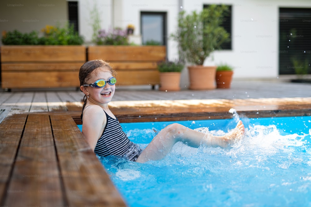 Une petite fille avec des lunettes à l’extérieur dans la cour, assise dans la piscine et regardant la caméra.