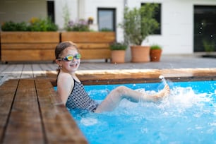 A small girl with goggles outdoors in the backyard, sitting in swimming pool and looking at camera.