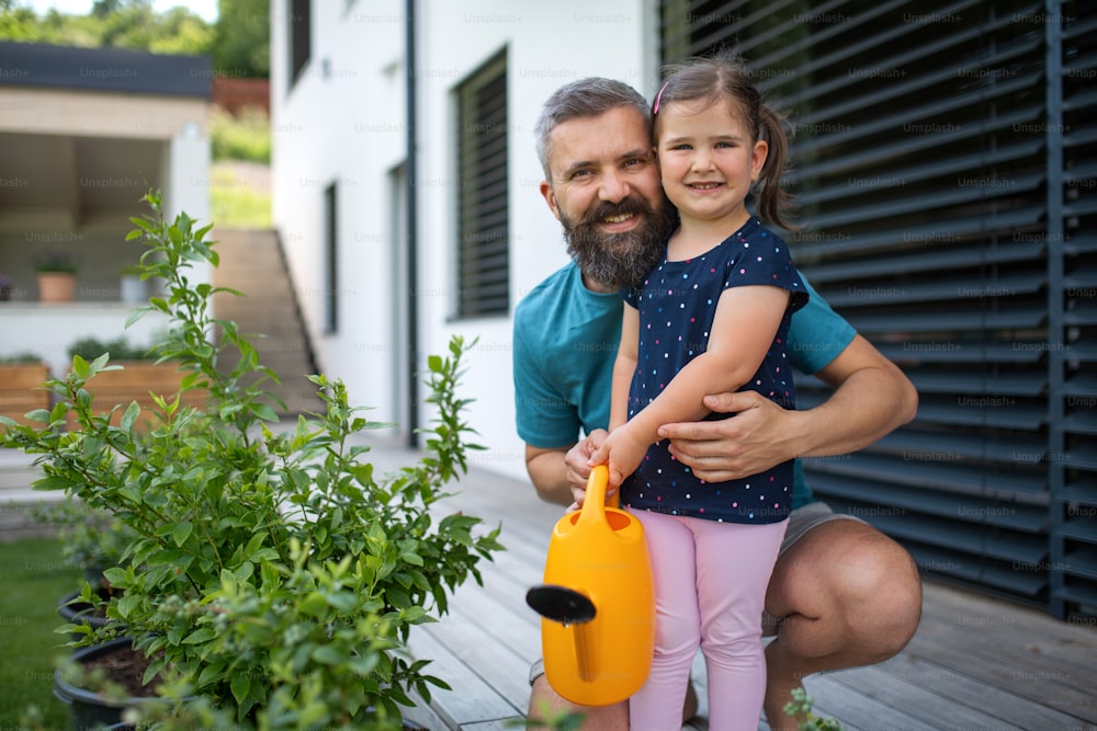 Un père et une petite fille avec arrosoir à l’extérieur dans la cour, regardant la caméra.
