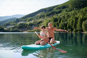 A happy preteen girl with grandfather on summer holiday by lake, paddleboarding.