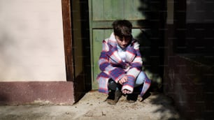 Una pobre niña triste con un gran abrigo al aire libre frente a la casa, concepto de pobreza.