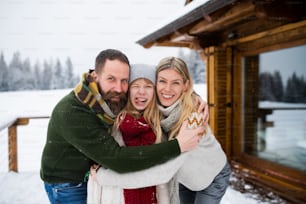 Portrait of a family with small daughter outdoors in winter holiday cottage, looking at camera.