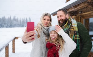 Uma família feliz com filha pequena tirando selfie na cabana de montanha ao ar livre no inverno.