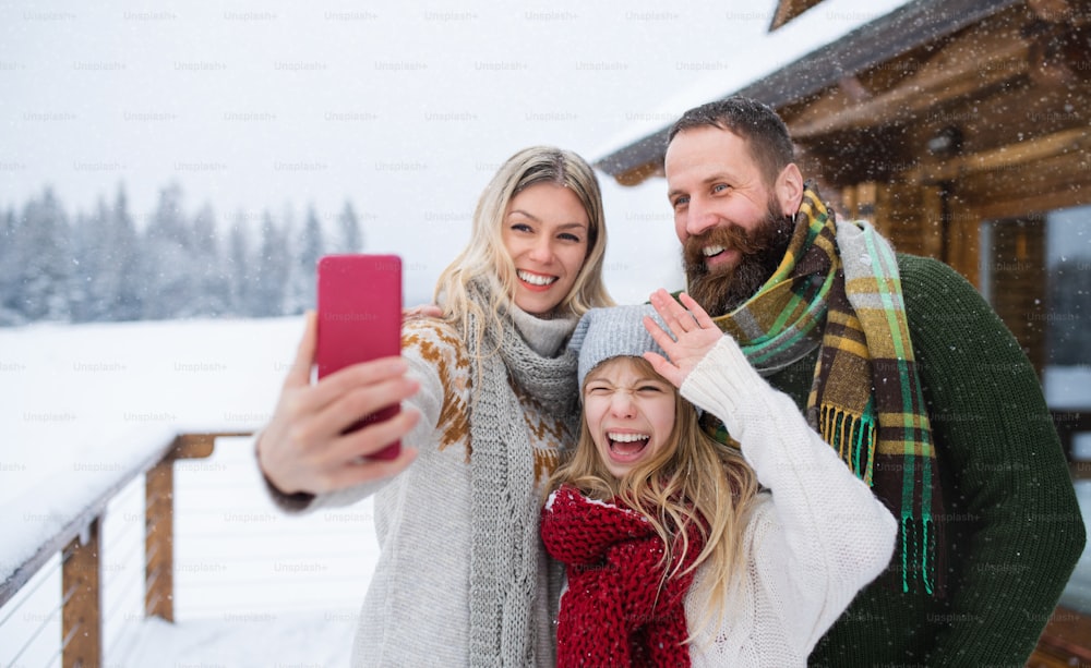 Una familia feliz con una hija pequeña que se toma una selfie junto a un refugio de montaña al aire libre en invierno.