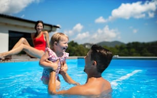 Happy young family with small daughter in swimming pool outdoors in backyard garden, playing.