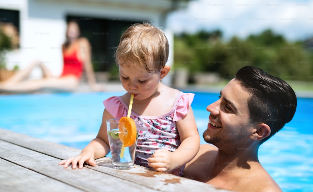 Portrait of small girl with father drinking lemonade in swimming pool outdoors in backyard garden.