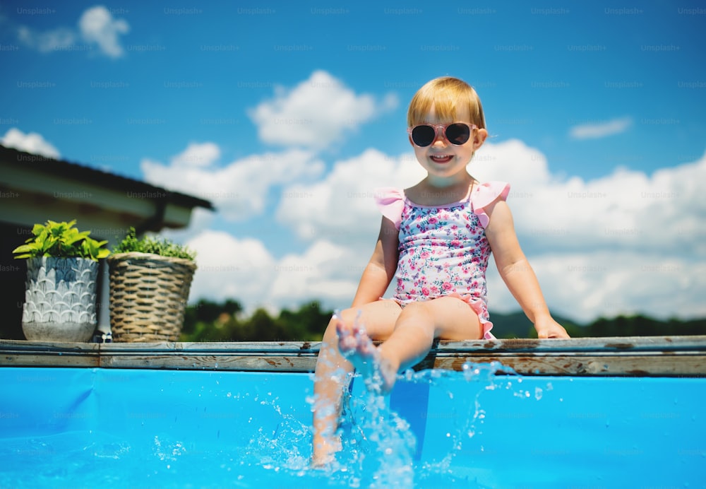 Retrato da menina pequena da criança sentada à beira da piscina ao ar livre no jardim do quintal, olhando para a câmera.