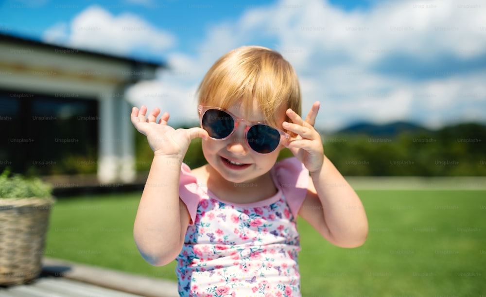 Retrato de una niña pequeña con gafas de sol al aire libre en el jardín del patio trasero, mirando a la cámara.