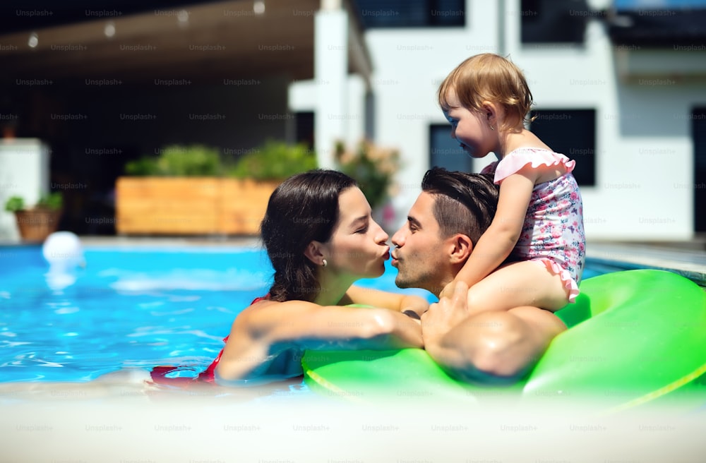 Happy young family with small daughter in swimming pool outdoors in backyard garden, kising.