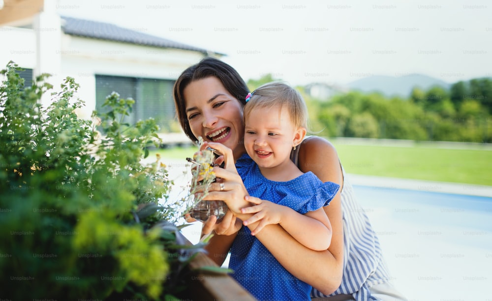Portrait d’une jeune mère avec une petite fille à l’extérieur dans le jardin arrière, pulvérisant des plantes.