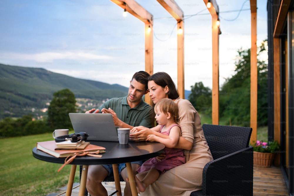 Familia joven con hija pequeña que usa computadora portátil al aire libre, fin de semana en casa de contenedores en el campo.