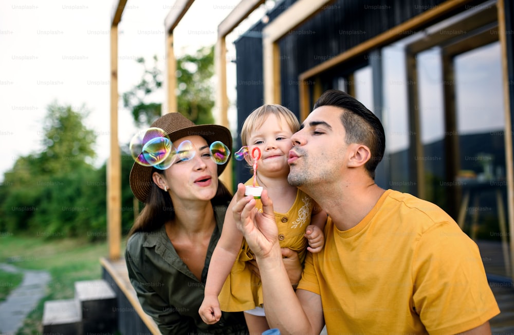 Familia joven con una hija pequeña soplando burbujas al aire libre, fin de semana en casa de contenedores en el campo.