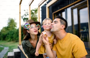 Giovane famiglia con figlia piccola che soffia bolle all'aperto, fine settimana in casa container in campagna.