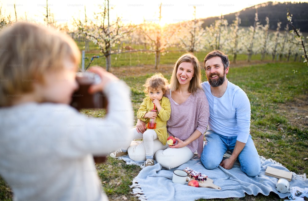 Small boy with camera taking photograph of family on picnic outdoors in spring nature at sunset.