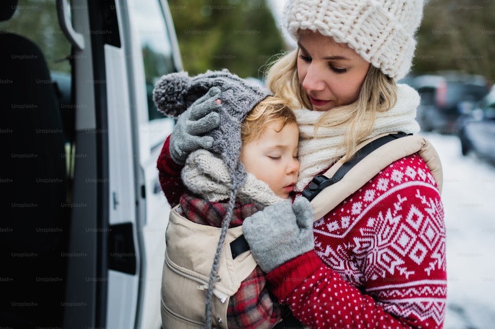 Portrait of mother with sleeping small daughter in carrier standing by car in winter nature.