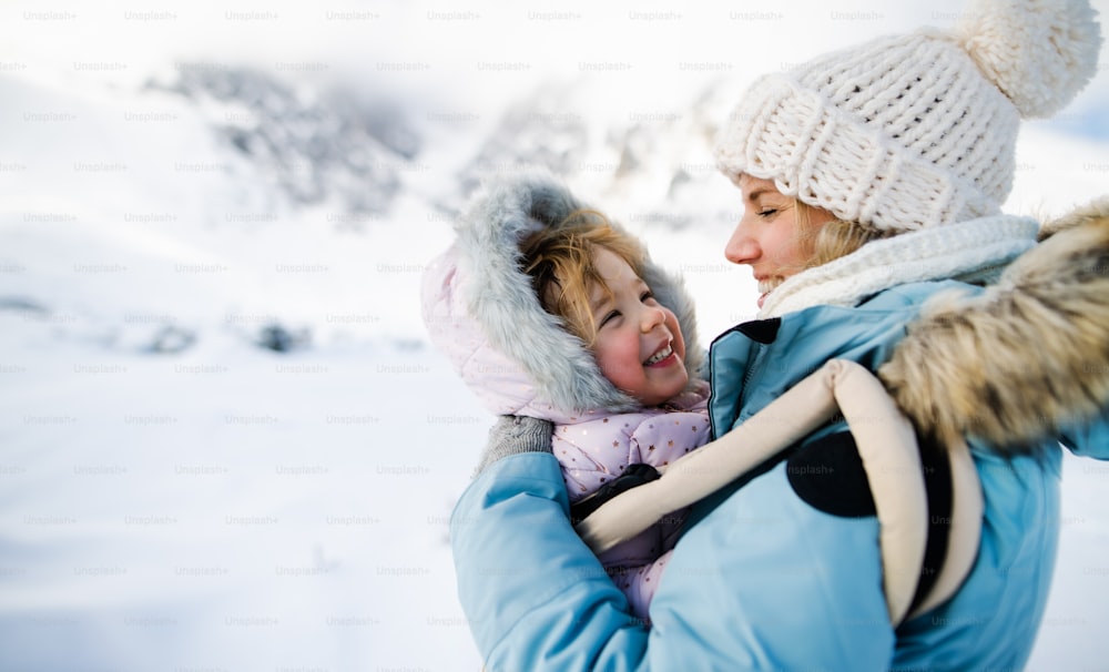 Portrait of mother with happy small daughter in carrier standing in winter nature, resting.