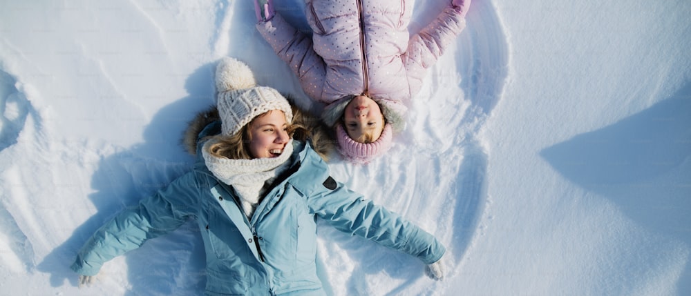 Top view portrait of cheerful mother with small toddler daughter lying in snow in winter nature.