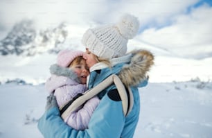 Portrait of mother with unhappy small daughter in carrier standing in winter nature, kissing.