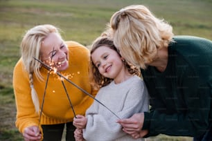 Happy small girl with mother and grandmother on a walk in nature, holding sparklers.