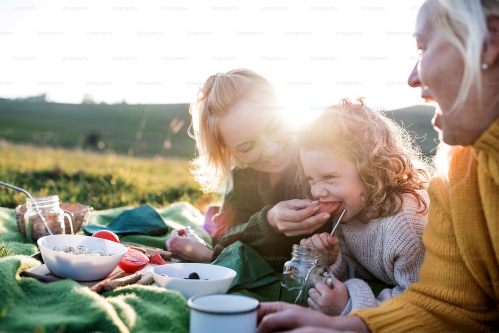 Happy small girl with mother and grandmother having picnic in nature at sunset.
