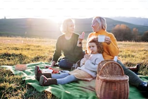 Happy small girl with mother and grandmother having picnic in nature at sunset.