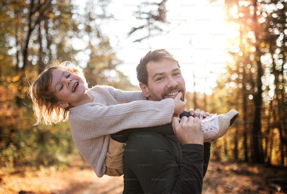 Mature father giving piggyback ride to happy small daughter on a walk in autumn forest.