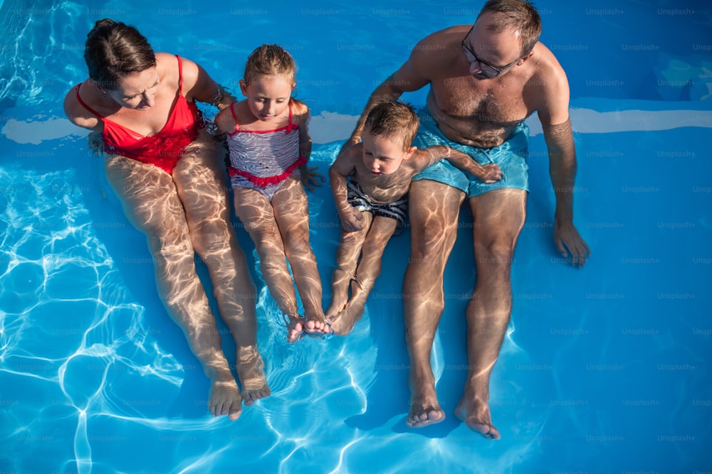 Young family with two small children sitting in swimming pool outdoors, top view.