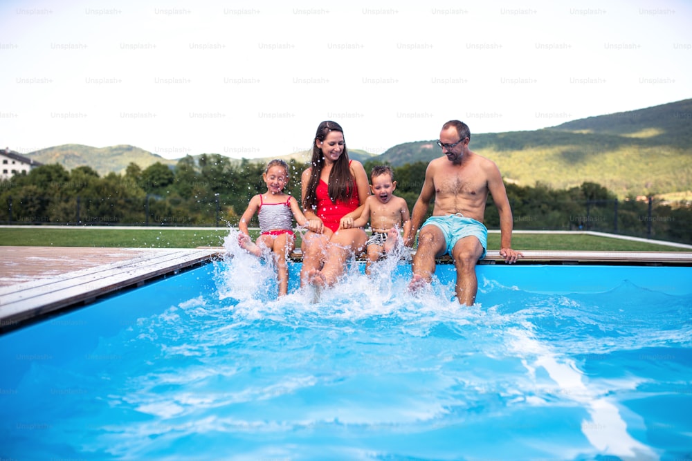Familia joven con dos niños pequeños sentados junto a la piscina al aire libre, divirtiéndose.