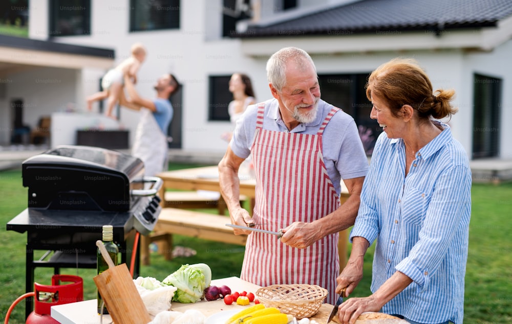 Vue de face d’un couple senior avec famille à l’extérieur sur barbecue de jardin, grillades.