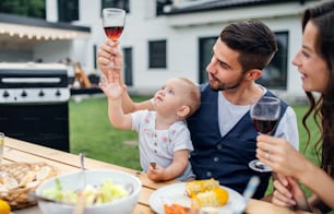 Giovane coppia con baby sitter al tavolo all'aperto sul barbecue del giardino di famiglia, bevendo vino.