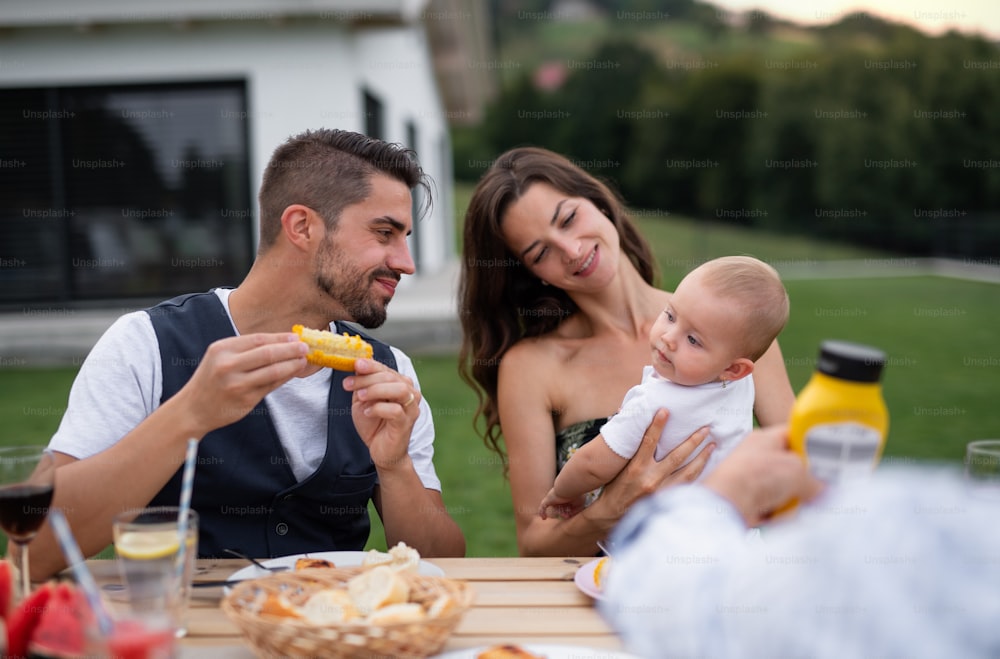 Pareja joven con bebé sentado en la mesa al aire libre en la barbacoa del jardín familiar, comiendo.