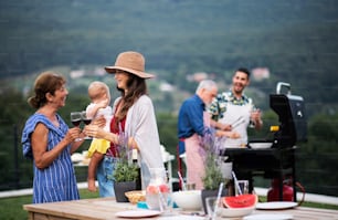 Famiglia multigenerazionale con vino all'aperto sul barbecue in giardino, grigliare e parlare.