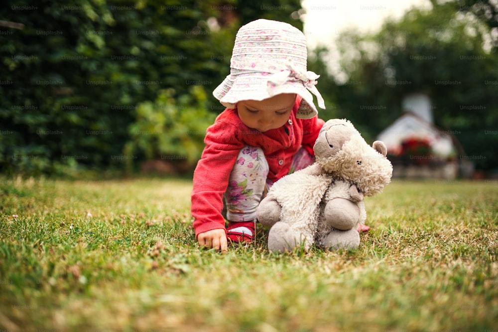 A front view of toddler girl standing outdoors on grass in garden in summer.