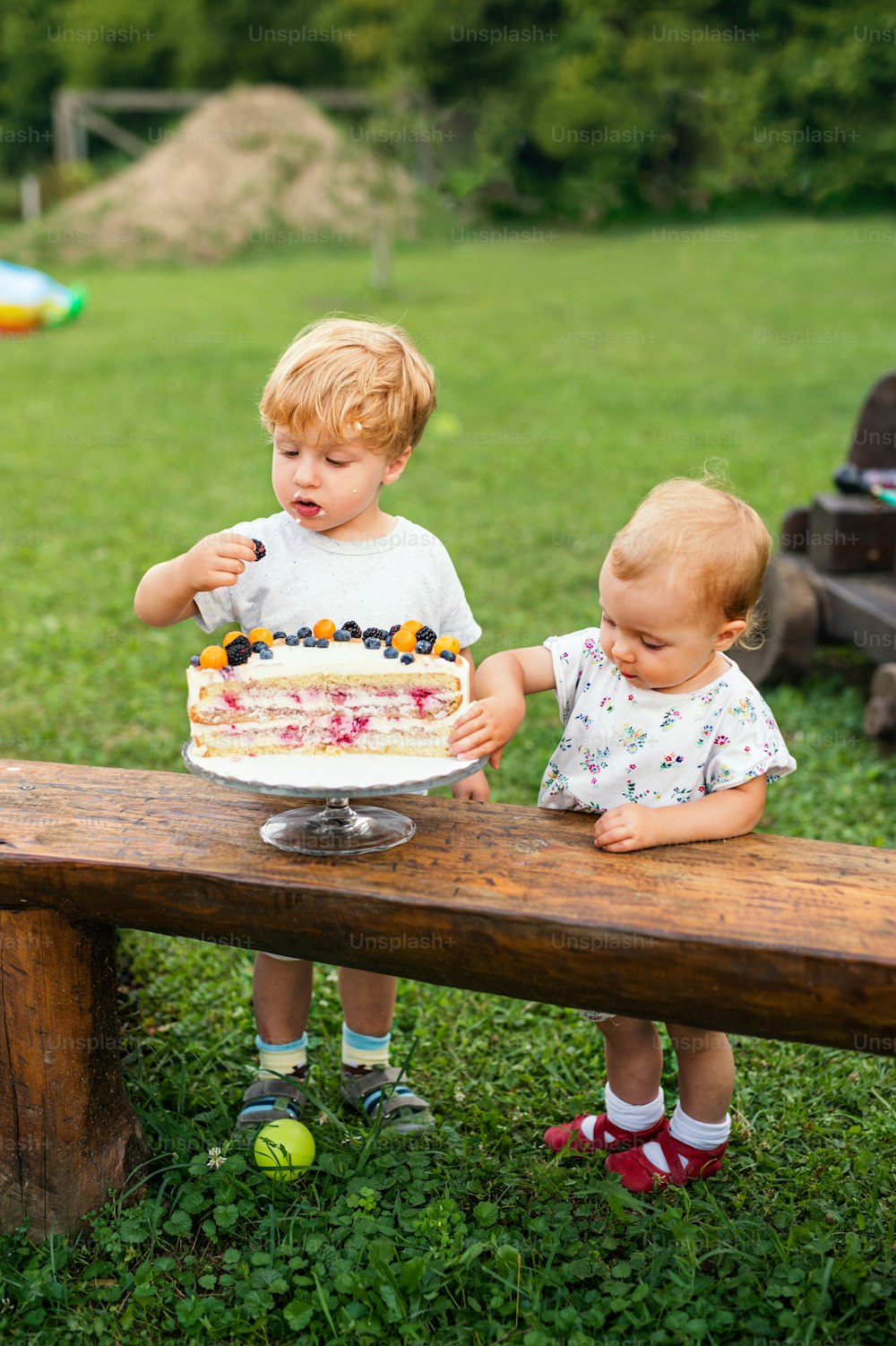 Front view of two toddler children with birthday cake outdoors in garden in summer.