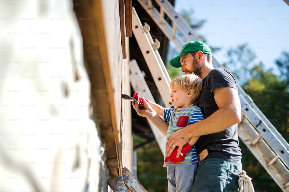 A side view of father and toddler boy outdoors in summer, painting wooden house.