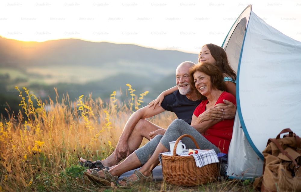 A small girl with grandparents sitting in shelter tent in nature at sunset, resting.