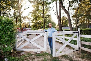 成熟した農家が家族の農場を屋外を歩き、門を開けるポートレート。